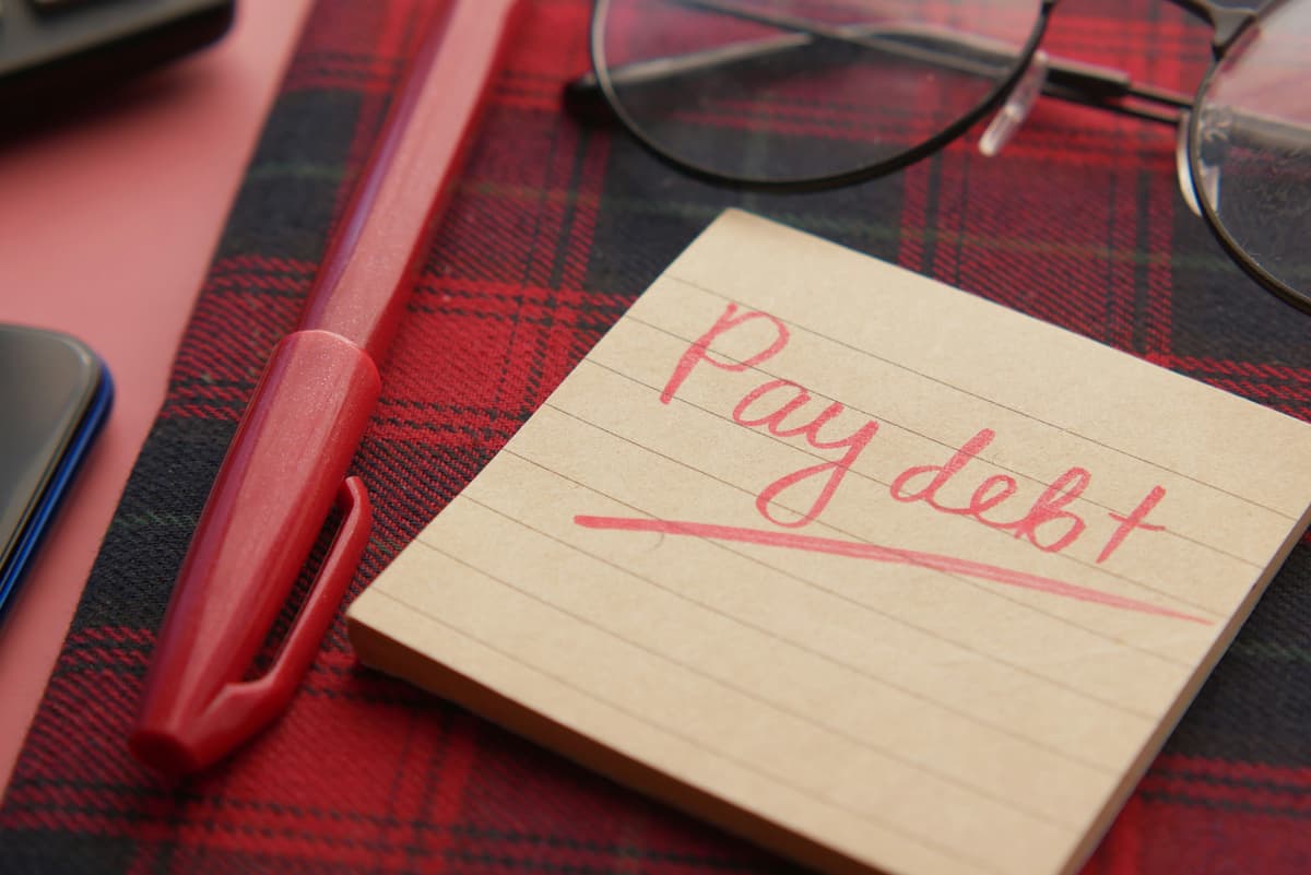 A handwritten note reading 'Pay Debt' on a notebook with a pen and glasses, highlighting financial planning and responsibility.