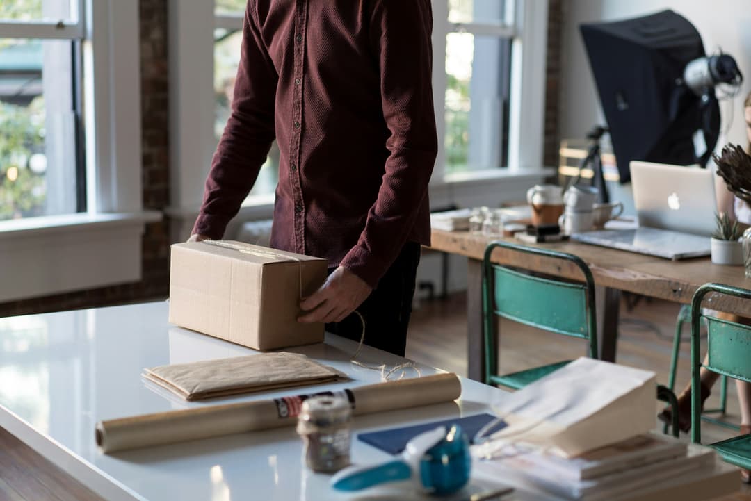 A person packaging a box on a white table in a cozy, well-lit workspace, symbolizing small business operations and e-commerce fulfillment.