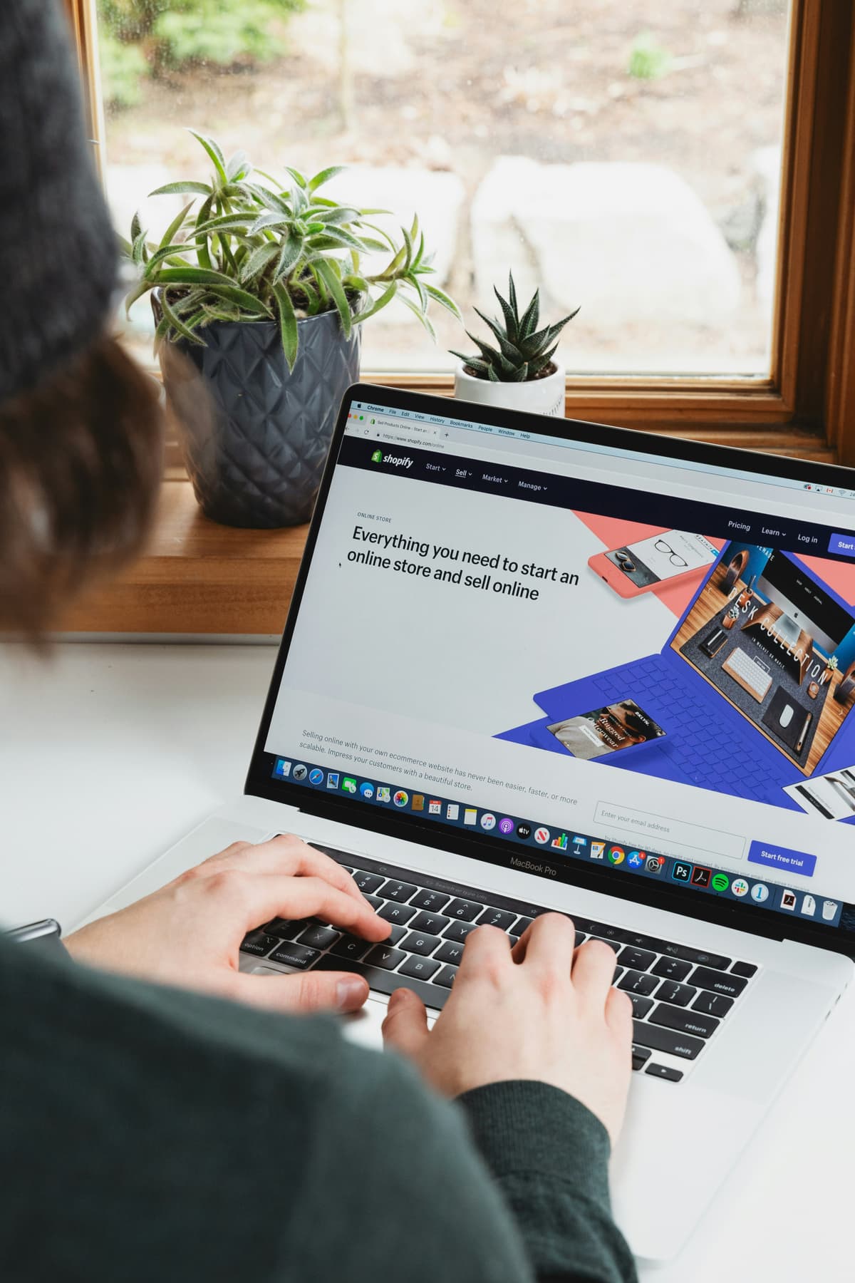 A person is working on a laptop with a high risk merchant account website open. The screen shows a colorful layout with images of digital devices and marketing tools. The laptop is placed on a wooden surface near a window, accompanied by potted plants in the background, providing a clean and natural workspace setting.