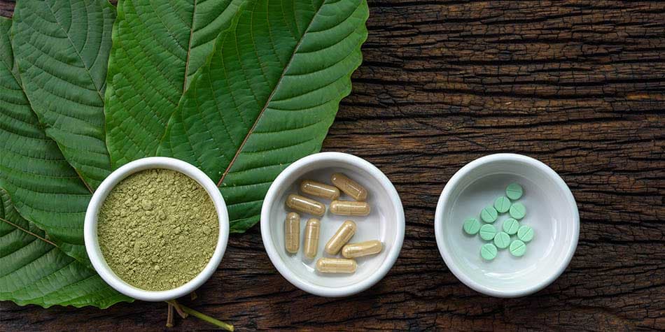 Green kratom powder and capsules displayed in small bowls alongside kratom leaves on a textured wooden surface.