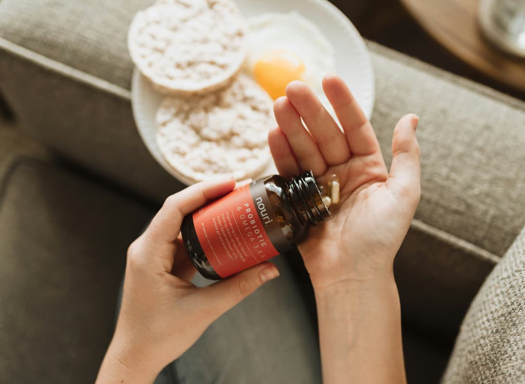 A close-up of a person holding a bottle of probiotics and omega supplements, with a plate of healthy breakfast items in the background.